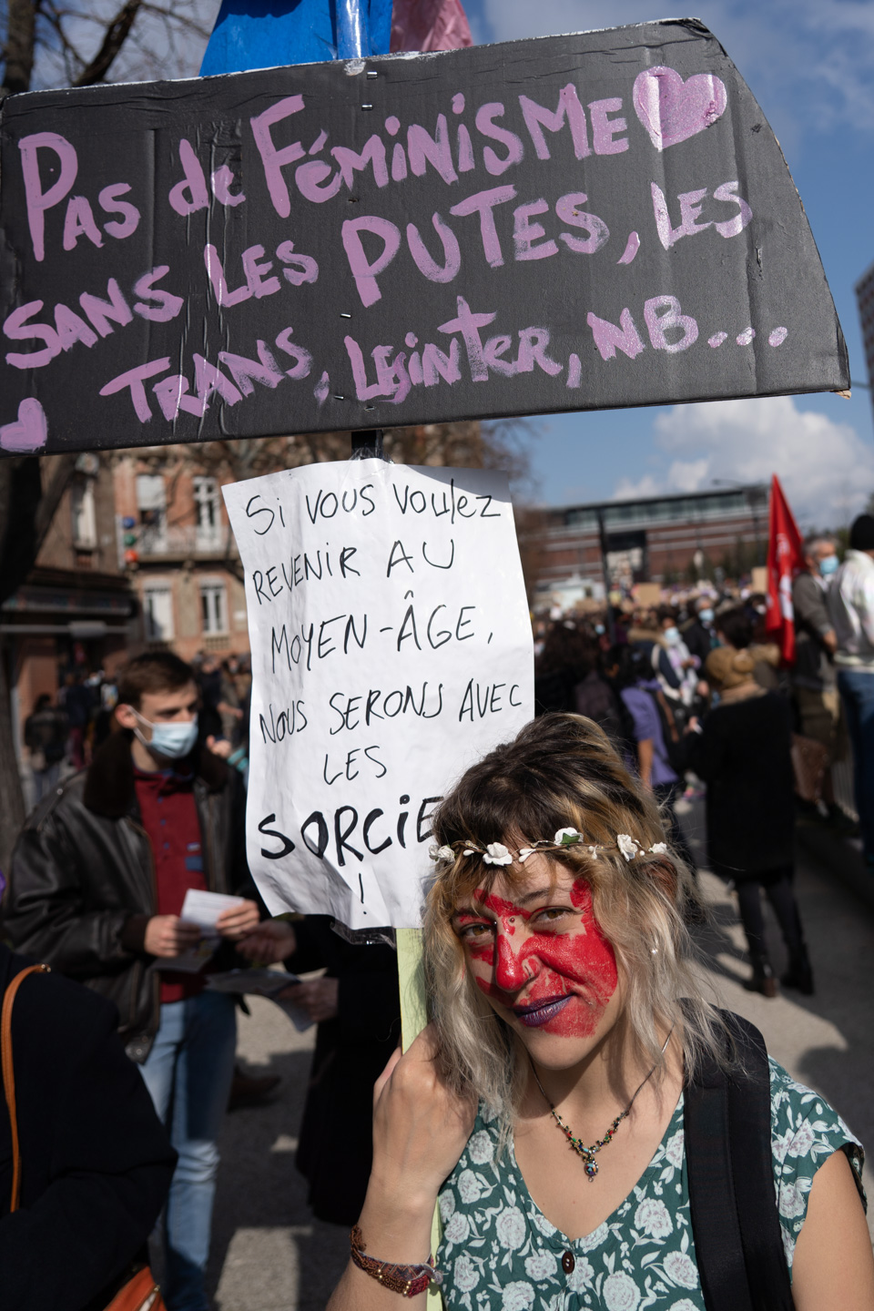 Une manifestante au visage peint en rouge, regard caméra, montre deux pancartes "Pas de féminisme sans les putes, les trans, les inter, nb..." et "Si vous voulez revenir au moyen-âge, nous serons avec les sorcières"  lors de la manifestation pour la journée sur les droits des femmes. Toulouse, 8 mars 2021.