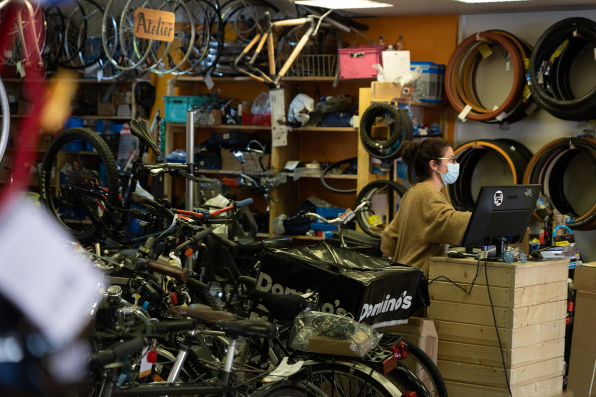 Vue sur l'intérieur d'une boutique de réparations de vélos. En premier plan, des vélos. En arrière plan, une employée masquée devant son ordinateur.