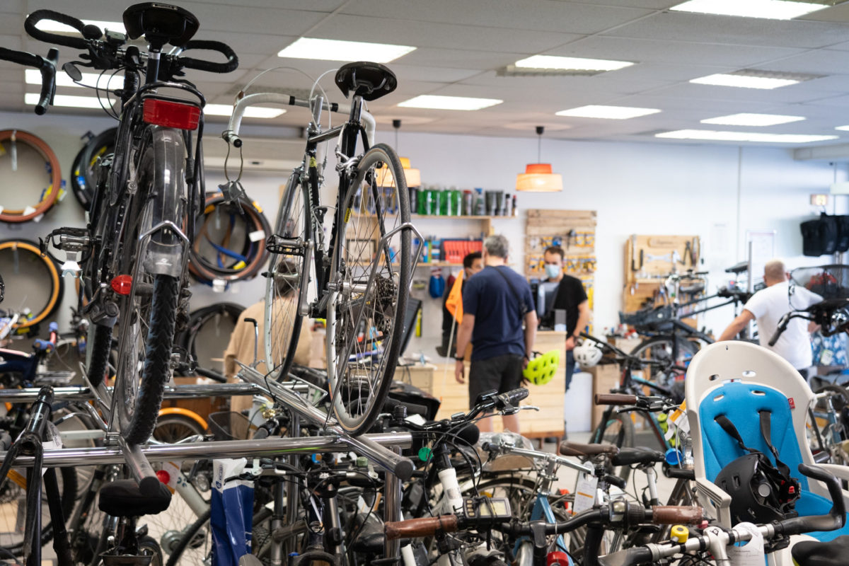 Vue sur l'intérieur d'une boutique de réparations de vélos. En premier plan, des vélos. En arrière plan, des clients.
